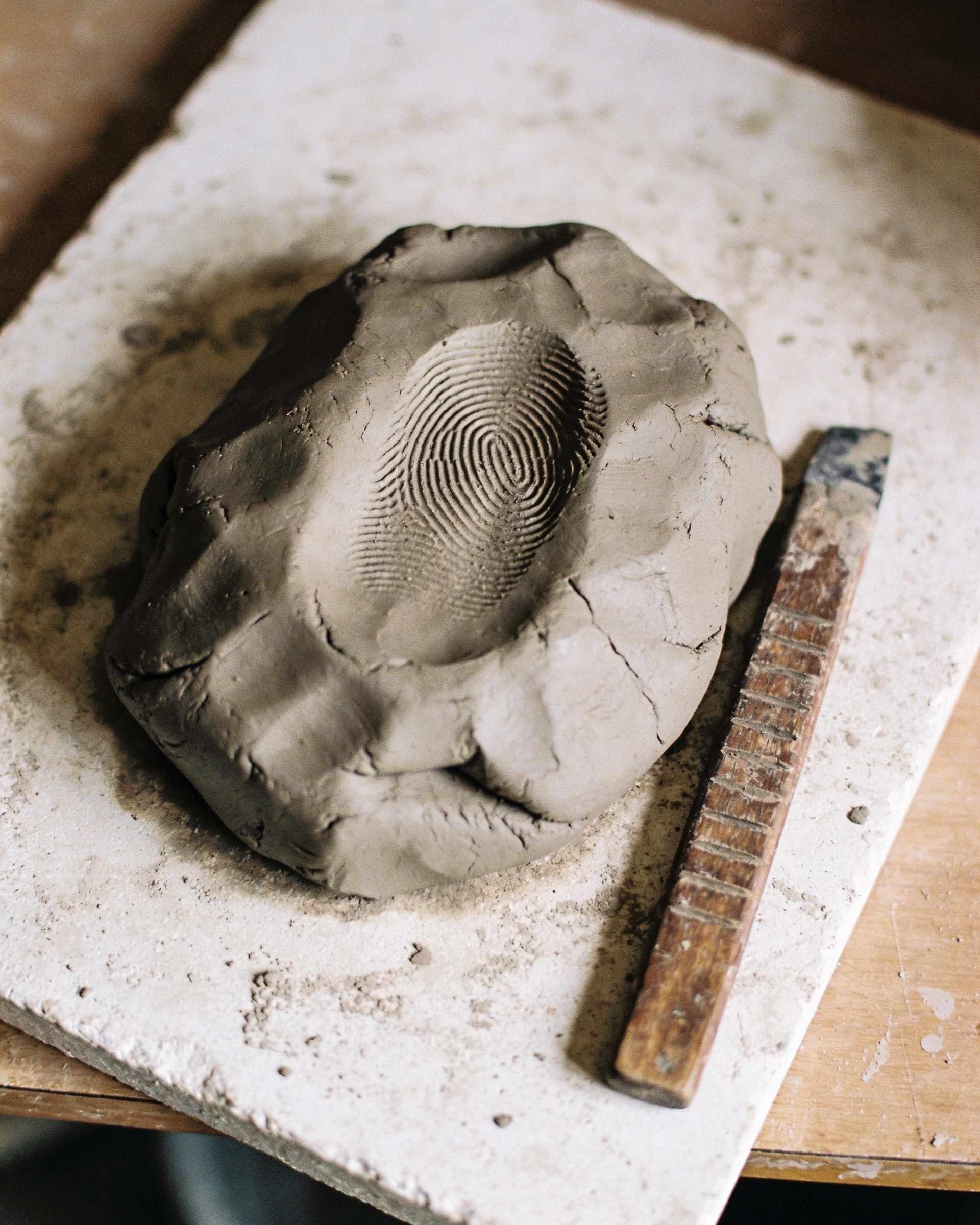 A lump of wet grey clay with a deep thumbprint and a worn wooden potter's rib on a dusty white plaster bat