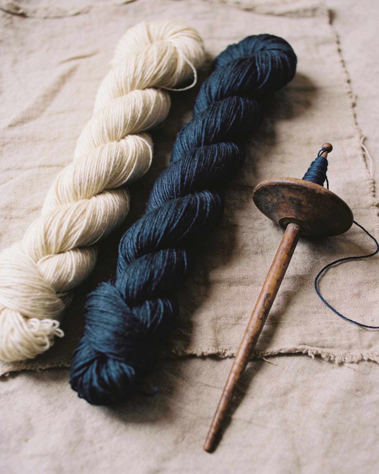 Skeins of undyed cream wool and deep indigo thread with an old wooden drop spindle on rough natural linen