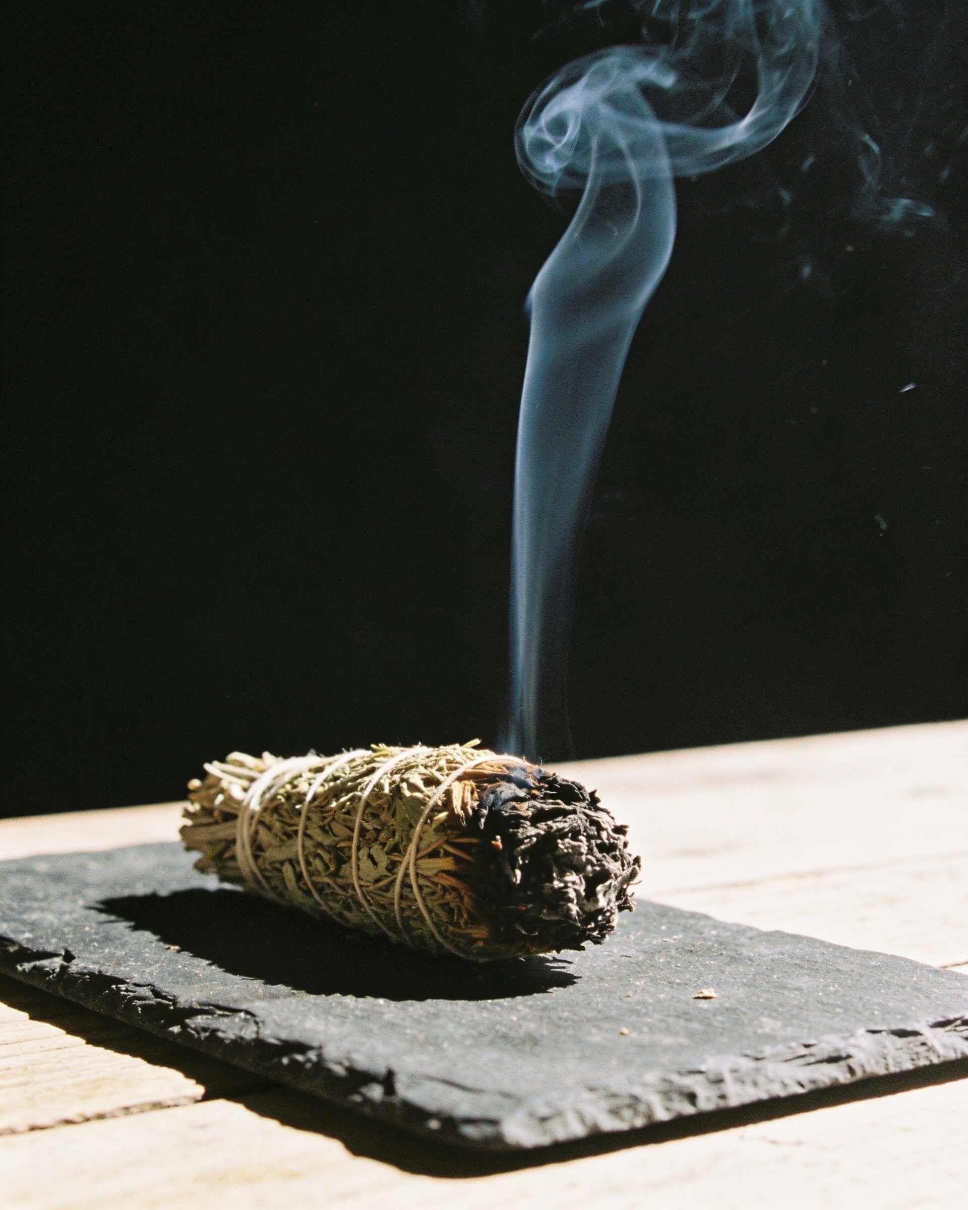 A thin curling plume of smoke rising from a smouldering bundle of dried herbs on a dark slate slab